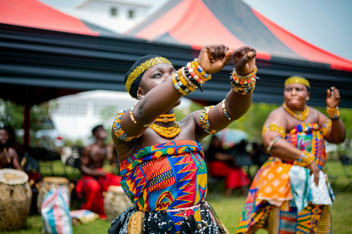 A dancer in kente cloth and beads, drummers behind her